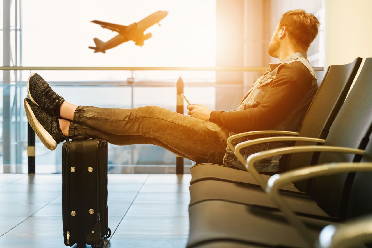 services-01 Side view full body of bearded man sitting on chair in waiting hall of airport and looking at flying aircraft behind window