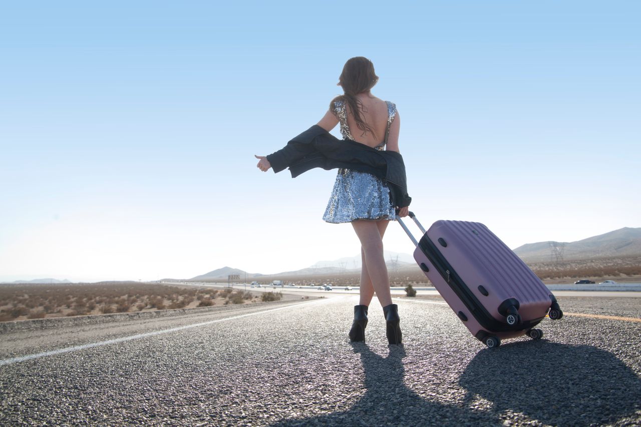 Home Woman in a sequined dress hitchhiking on a desert road with a pink suitcase. Fashion meets adventure.